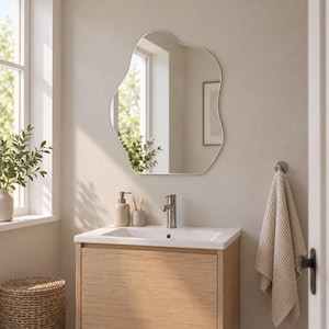 May include: An irregular, organic-shaped wall mirror with a thin frame hangs above a bathroom vanity. The vanity features a light wood cabinet, a white rectangular sink, a silver tap, a soap dispenser, and a toothbrush holder.