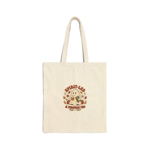 Può includere: Una borsa in tela color crema con una grafica centrale raffigurante un fantasma che tiene in mano una tazza di caffè e un libro, circondato da zucche e foglie autunnali. Il testo "SPIRIT-LED & PUMPKIN FED" è stampato in marrone.