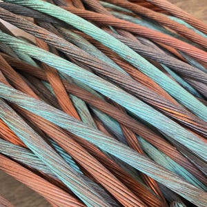 Może przedstawiać: Zbiór splecionych metalowych kabli z mieszanką odcieni utlenionej miedzi, w tym jasnego metalicznego pomarańczu, ciemnego brązu i żywej turkusowej zieleni. Sploty są ciasno skręcone w grube, teksturowane liny druciane.