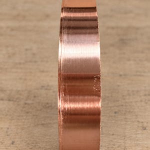 May include: A solid copper ring with a smooth, polished finish and a flat, wide band profile. The metal surface displays a warm, metallic reddish-brown hue with subtle vertical light reflections along the curved outer edge of the band.