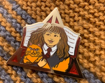 May include: A gold-colored enamel pin featuring a cartoon illustration of a character with brown hair, a red tie, and a cat. The pin is shaped like a triangle with a book and the character's image inside. The background is a knitted fabric.