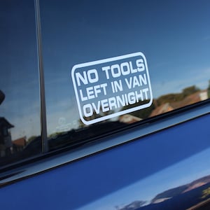 May include: A white rectangular vinyl decal applied to a dark blue vehicle window. The decal features a rounded border and the text in white capital letters reads: NO TOOLS LEFT IN VAN OVERNIGHT. The sticker is positioned on the glass surface.