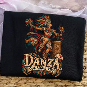 May include: A black t-shirt featuring a colourful graphic of a dancer in traditional regalia with a large feathered headdress, holding fans, standing next to a carved drum. The text reads DANZA QUE SIGUE VIVA in a decorative, weathered font.