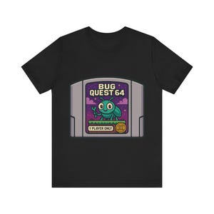 May include: A black t-shirt featuring a graphic of a gray video game cartridge. The label displays the text BUG QUEST 64, a cartoon teal bug holding a wrench, a purple sky with stars, a green grassy platform, and the text 1 PLAYER ONLY.