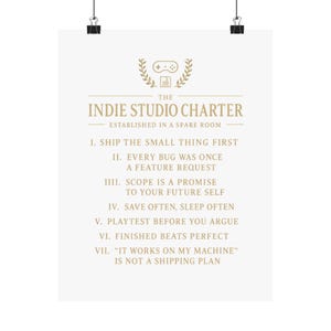 Puede incluir: Un póster blanco con el texto dorado The Indie Studio Charter Established in a Spare Room, que incluye siete reglas numeradas como Ship the Small Thing First, Every Bug Was Once a Feature Request e It Works on My Machine is Not a Shipping Plan.