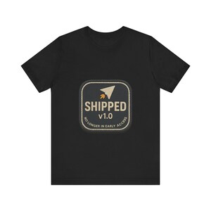 Könnte beinhalten: Ein schwarzes Kurzarm-T-Shirt mit einem mittigen, quadratischen Aufnäher mit abgerundeten Ecken. Der Aufnäher zeigt ein beiges Papierflieger-Symbol, den Text SHIPPED v1.0 und den Schriftzug NO LONGER IN EARLY ACCESS in beiger Farbe.