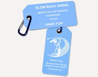 May include: Two light blue rectangular tags attached to a dark blue carabiner. The top tag features white text: SLOW BACK SWING, SMOOTH FOLLOW THROUGH, CONFIDENCE, FOCUS, HAVE FUN. The bottom tag shows a white golfer graphic and text: Mindset Tags, Because Every Swing Begins in Your Mind, Mindsettags.com.