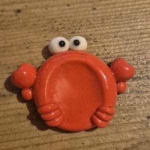 Op de afbeelding: Een feloranje, cirkelvormig object in de vorm van een krab met een gladde, glanzende afwerking. Het heeft twee opvallende witte, bolvormige ogen met zwarte pupillen aan de bovenkant, twee omhoogstaande scharen aan de zijkanten en vier kleine pootjes.