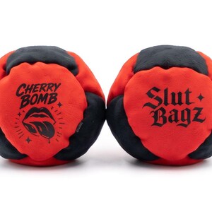 May include: Two round, multi-paneled bean bags featuring a red and black color scheme. The left bag displays the text "CHERRY BOMB" above a graphic of lips with a tongue. The right bag features the text "Slut Bagz" in a gothic-style font.