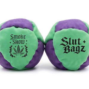 May include: Two round, multi-paneled footbags made of purple and light green fabric. The left footbag features black text reading Smoke Show with a cannabis leaf graphic. The right footbag features black text reading Slut Bagz in gothic font.