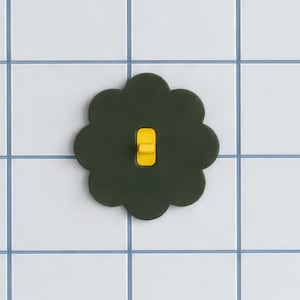 Op de afbeelding: Een donkergroene, bloemvormige afdekplaat voor een lichtschakelaar met een rechthoekige uitsparing in het midden. Een felgele tuimelschakelaar bevindt zich in het midden van de donkergroene plaat, contrasterend met de matte afwerking van de vorm.