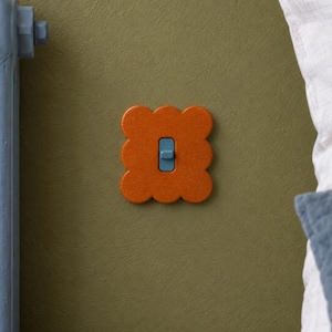 Op de afbeelding: Een lichtschakelaarafdekplaat met een geschulpte, wolkachtige vierkante vorm in een levendige oranje kleur. Het midden is voorzien van een rechthoekige blauwe tuimelschakelaar. De plaat is gemonteerd op een gestructureerde olijfgroene muur.