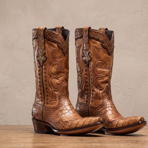 May include: A pair of tan leather cowboy boots featuring a textured crocodile-skin pattern on the foot and toe, with intricate decorative stitching and embossed details on the tall shaft, finished with a stacked heel and a pointed toe shape.