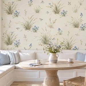 May include: Wallpaper featuring a repeating pattern of green grasses, blue wildflowers, and small birds on a cream background. The scene includes a white built-in bench with blue and white striped pillows, a round wooden table, and a ceramic pitcher.