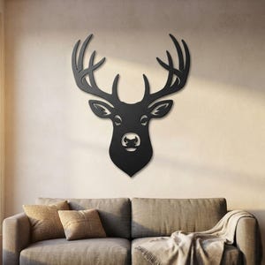 Puede incluir: Una pieza de arte mural de metal negro mate con forma de cabeza y cornamenta de ciervo. La silueta presenta detalles recortados para los ojos, las orejas y la nariz, creando una representación plana y estilizada de un ciervo en la pared.