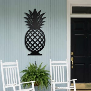 Op de afbeelding: Een matzwart metalen wanddecoratie in de vorm van een ananas met een gedetailleerd ruitpatroon op het vruchtlichaam en puntige bladeren aan de bovenkant. Het silhouet is plat en heeft een symmetrisch ontwerp met een stevige basis en top.