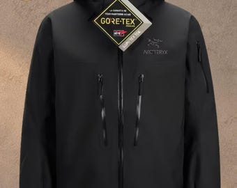 May include: A black Arc'teryx hooded jacket featuring a full-length front zip, two vertical chest pockets with black zips, and a Gore-Tex tag attached to the collar. The brand logo is embroidered in grey on the upper left chest area.