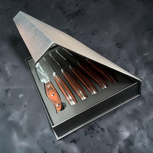 May include: A set of five steak knives with polished metal blades and dark brown wood grain handles, arranged in a gray foam-lined tray. The set is housed in a triangular, metallic-finished box with a hinged lid that is partially open.