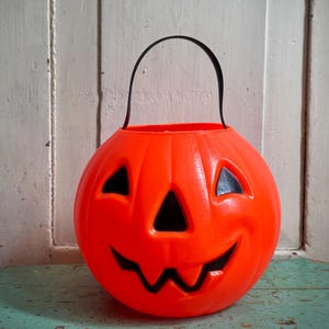 May include: A bright orange plastic pumpkin bucket featuring a carved jack-o'-lantern face with black triangular eyes, a triangular nose, and a jagged mouth. The container includes a thin, flexible black plastic handle attached to the top rim.