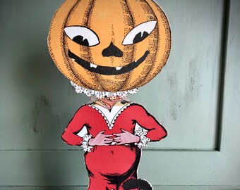 May include: A vintage Halloween cardboard cutout of a pumpkin-headed figure wearing a red costume and holding a black cat. The figure is standing on a wooden floor.