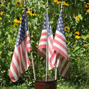 May include: Three American flags with red, white, and blue stripes and a blue canton with white stars are mounted on wooden poles with gold-toned finials. The flags are held upright in a weathered, cylindrical red metal container.
