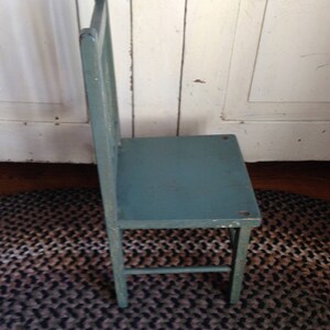 Puede incluir: Una silla de madera azul verdosa con un dise&ntilde;o sencillo. La silla tiene un respaldo de listones y un asiento &uacute;nico y ancho. La silla est&aacute; sentada sobre una alfombra trenzada marr&oacute;n y negra.