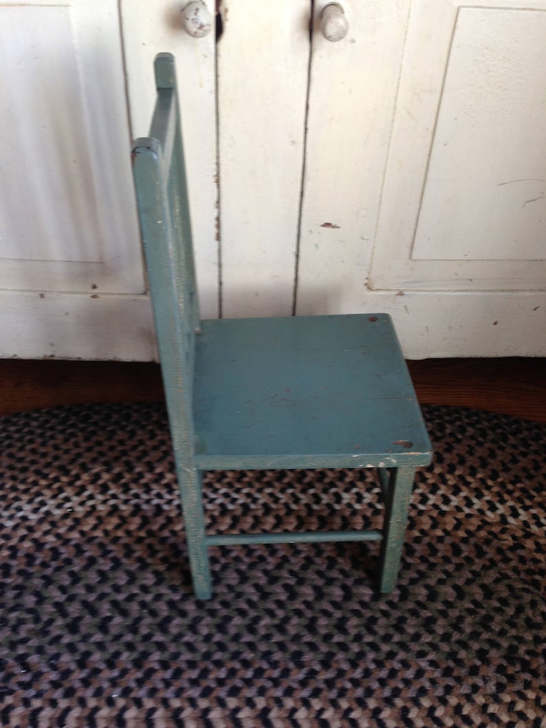 Puede incluir: Una silla de madera azul verdosa con un dise&ntilde;o sencillo. La silla tiene un respaldo de listones y un asiento &uacute;nico y ancho. La silla est&aacute; sentada sobre una alfombra trenzada marr&oacute;n y negra.