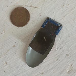 May include: A small, vintage metal clip with a blue painted top section featuring the embossed text KIRCHHOF. The lower portion is a smooth, light gray curved metal scoop shape. A copper one-cent coin is positioned next to the clip for scale.
