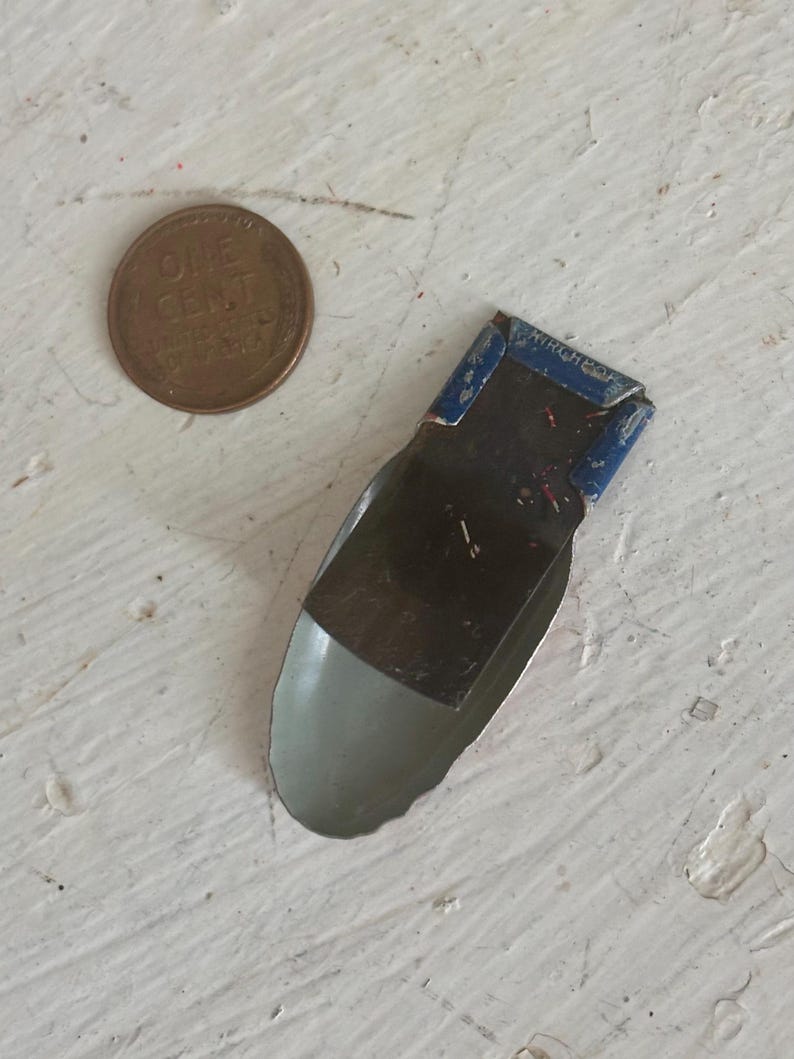 May include: A small, vintage metal clip with a blue painted top section featuring the embossed text KIRCHHOF. The lower portion is a smooth, light gray curved metal scoop shape. A copper one-cent coin is positioned next to the clip for scale.