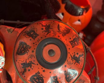 May include: A vintage Halloween-themed tin lid with a central black circle and a surrounding orange field. The lid features black illustrations of witches, pumpkins, owls, and stars. The lid is surrounded by other orange Halloween decorations.