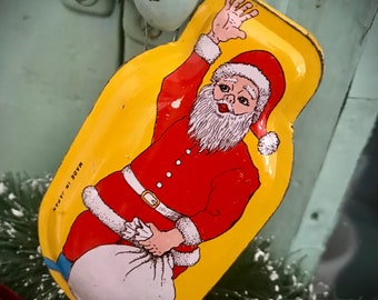 May include: A vintage tin ornament of Santa Claus holding a bag of gifts. He is wearing a red suit with white trim and a white beard. The ornament is yellow with the words "Made in Japan" printed on the back.