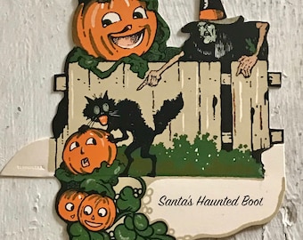 Könnte beinhalten: Eine Vintage-Halloween-Karte mit einer Hexe, einer schwarzen Katze und Kürbissen. Die Hexe trägt einen schwarzen Hut und ein schwarzes Gewand und schaut über einen Holzzaun. Die schwarze Katze sitzt neben einem Kürbis und schaut die Hexe an. Die Karte trägt den Text "Santa's Haunted Boot".