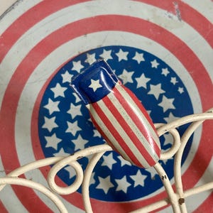 May include: A small, metal, bomb-shaped object featuring a red, white, and blue striped pattern with a single white star on a blue field. It is positioned on a cream-colored wire rack in front of a circular tin with a red, white, and blue star design.