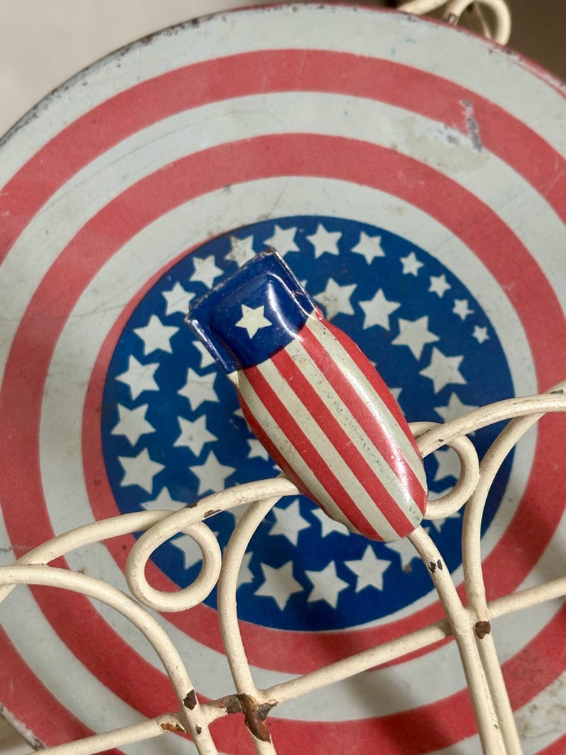 May include: A small, metal, bomb-shaped object featuring a red, white, and blue striped pattern with a single white star on a blue field. It is positioned on a cream-colored wire rack in front of a circular tin with a red, white, and blue star design.