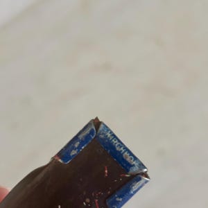May include: A dark brown, rectangular object with a blue metal cap on the end. The blue cap features the embossed text "KIRCHHOF" in capital letters. The surface of the brown section shows small, scattered red and white flecks of debris.
