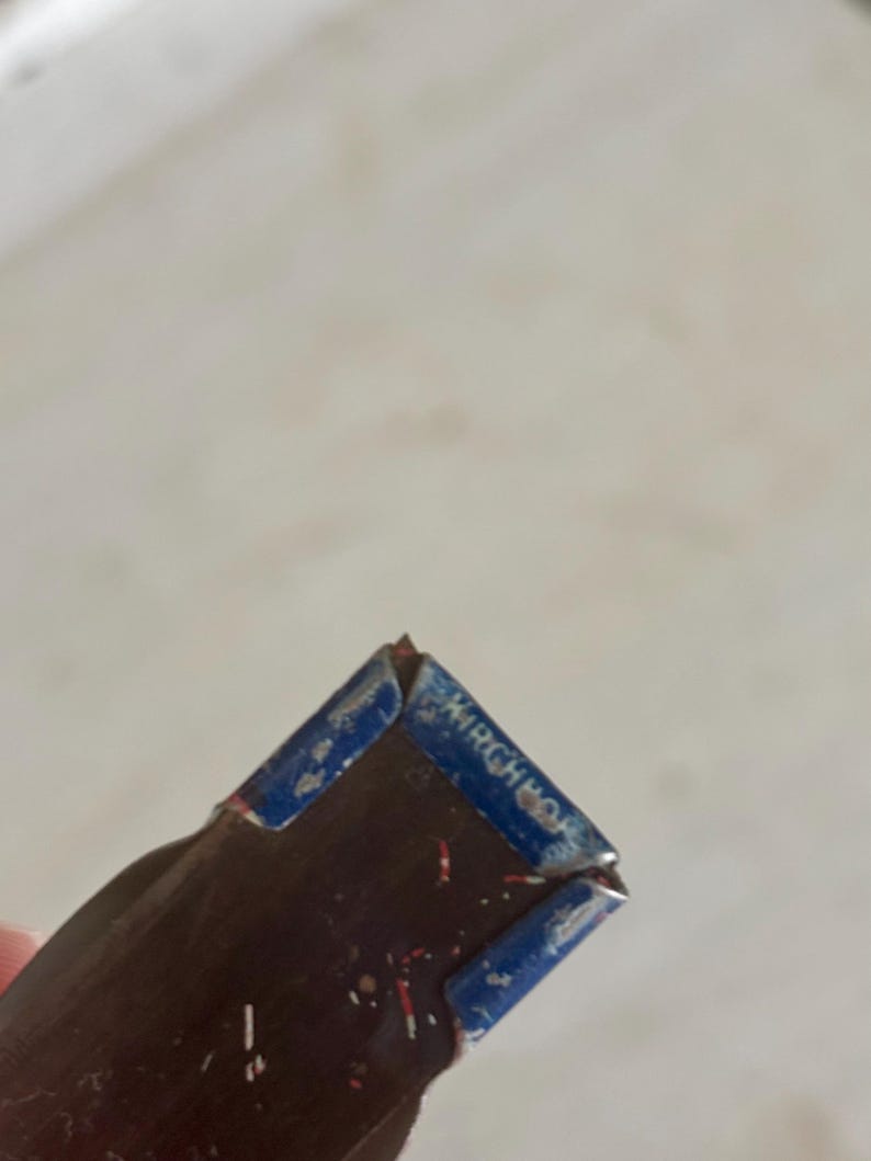 May include: A dark brown, rectangular object with a blue metal cap on the end. The blue cap features the embossed text "KIRCHHOF" in capital letters. The surface of the brown section shows small, scattered red and white flecks of debris.