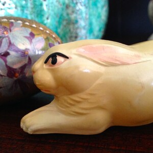 May include: A white ceramic rabbit figurine with a pink ear and black eye. The rabbit is lying on its side with its head turned to the side.