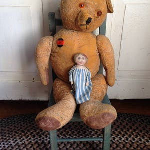 Puede incluir: Un gran oso de peluche desgastado de color marr&oacute;n claro con ojos rojos est&aacute; sentado en una silla de madera verde. Una peque&ntilde;a mu&ntilde;eca de porcelana con un vestido a rayas azules y blancas est&aacute; sentada en el regazo del oso. El oso tiene un bot&oacute;n rojo en el pecho que dice "Trick or Treat".