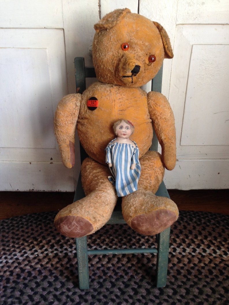 Puede incluir: Un gran oso de peluche desgastado de color marr&oacute;n claro con ojos rojos est&aacute; sentado en una silla de madera verde. Una peque&ntilde;a mu&ntilde;eca de porcelana con un vestido a rayas azules y blancas est&aacute; sentada en el regazo del oso. El oso tiene un bot&oacute;n rojo en el pecho que dice "Trick or Treat".