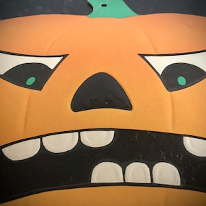 May include: A close-up of a carved pumpkin with a scary face. The pumpkin has a wide, toothy grin, green eyes, and a black nose. The pumpkin is orange and has a green stem.