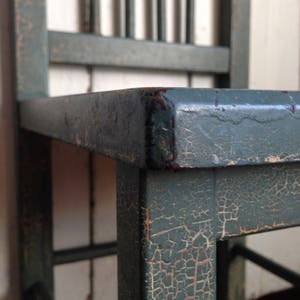 Puede incluir: Primer plano de una silla de madera verde con pintura agrietada y astillada. La silla tiene un dise&ntilde;o sencillo con listones verticales en el respaldo y un asiento cuadrado.