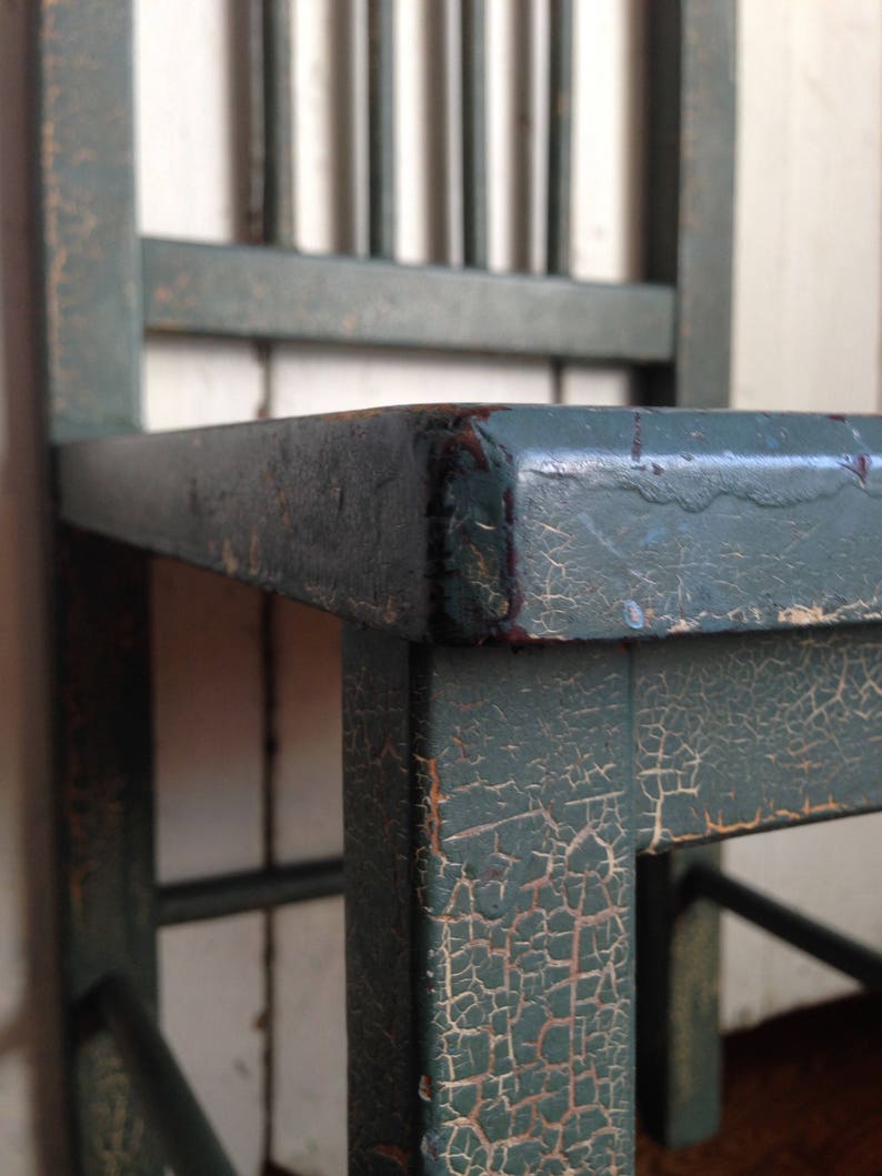 Puede incluir: Primer plano de una silla de madera verde con pintura agrietada y astillada. La silla tiene un dise&ntilde;o sencillo con listones verticales en el respaldo y un asiento cuadrado.