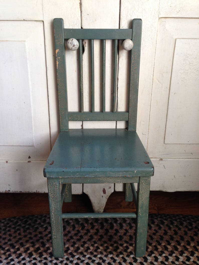 Puede incluir: Una silla infantil vintage pintada de verde con pomos blancos en el respaldo. La silla tiene un acabado desgastado y est&aacute; sobre una alfombra con estampado de blanco y negro.