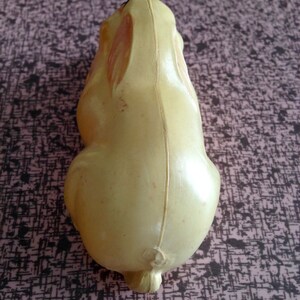 May include: A yellow plastic toy rabbit with a brown nose and black eyes. The rabbit is sitting on a pink and black patterned surface.