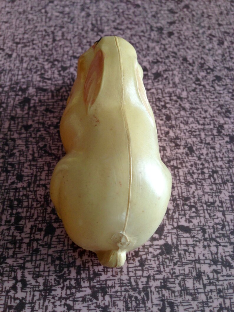 May include: A yellow plastic toy rabbit with a brown nose and black eyes. The rabbit is sitting on a pink and black patterned surface.