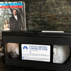 May include: A VHS tape of the movie "Captain Kronos: Vampire Hunter". The tape is black with a white label that says "Captain Kronos: Vampire Hunter" in bold letters. The label also includes the movie's rating, "R", and the runtime, "1074 Color 91 Min".