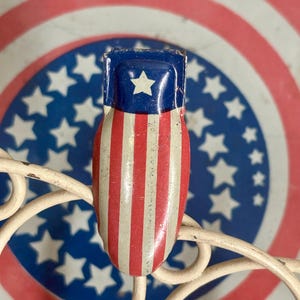 May include: A metal clip featuring a patriotic design with a blue top section containing a single white star, followed by vertical red and white stripes. The clip is positioned against a circular background with concentric red and white rings.