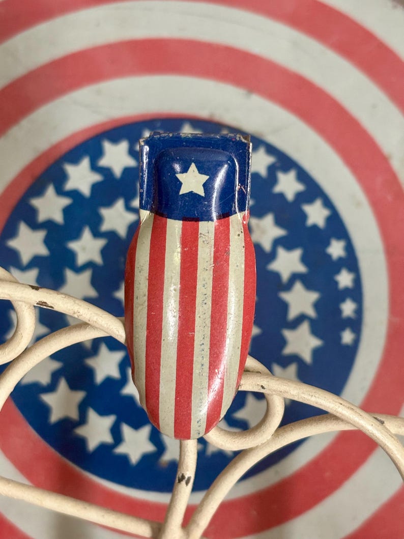 May include: A metal clip featuring a patriotic design with a blue top section containing a single white star, followed by vertical red and white stripes. The clip is positioned against a circular background with concentric red and white rings.