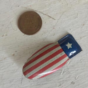 May include: A small, curved metal pin featuring a red, white, and blue flag design with a single white star on a blue field and red and white stripes. A copper one-cent coin is positioned next to the pin for scale, showing its small size.