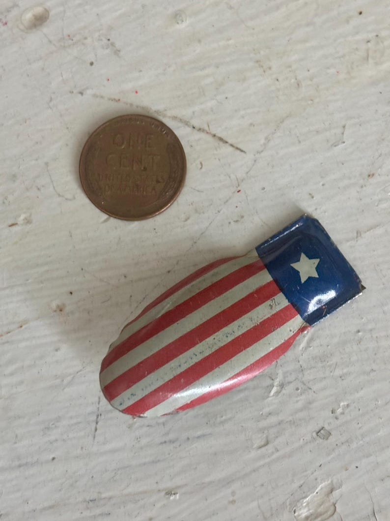 May include: A small, curved metal pin featuring a red, white, and blue flag design with a single white star on a blue field and red and white stripes. A copper one-cent coin is positioned next to the pin for scale, showing its small size.